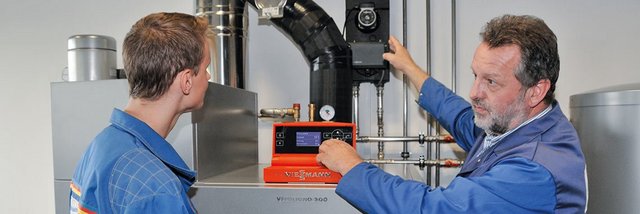 Der ältere Mann zeigt dem jüngeren Techniker, wie man ein Steuergerät an einer Heizungsanlage bedient.