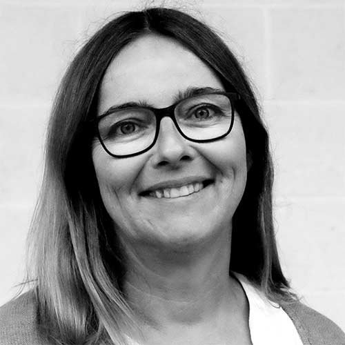 In einem hellen Raum lächelt eine Frau mit Brille, ihre Haare sind leicht gewellt und sie trägt ein schlichtes Oberteil.