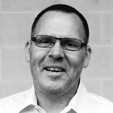 Der Mann zeigt ein freundliches Lächeln, während er in die Kamera schaut, umgeben von einer schlichten, grauen Kulisse.