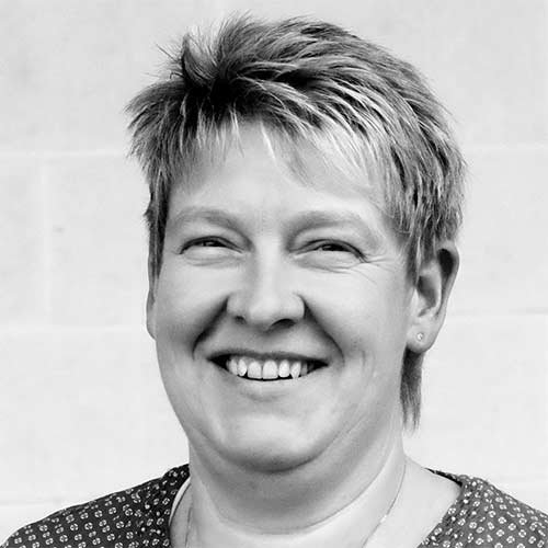 Die Frau mit fröhlichem Gesichtsausdruck hat kurzes, helles Haar und trägt ein gemustertes Shirt.