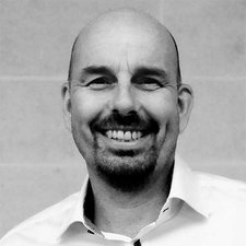 Der Mann hat ein breites Lächeln und trägt ein Hemd, während er vor einer neutralen Wand steht.