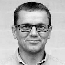 Der Mann hat eine entspannte Ausstrahlung, mit einem leichten Lächeln und einem neutralen Hintergrund in Grautönen.